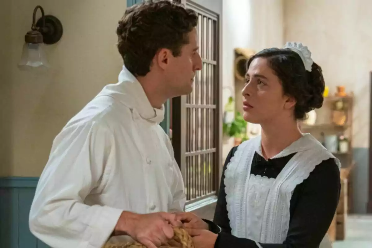 Fotografía de Lope vestido de chef y Vera vestida de sirvienta se miran fijamente en una habitación con decoración antigua en La Promesa