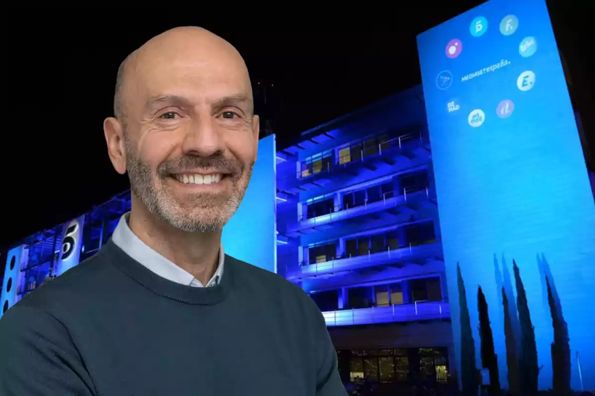 Alberto Carullo somrient amb barba i suèter fosc davant de l'edifici de Mediaset España il·luminat de blau a la nit.