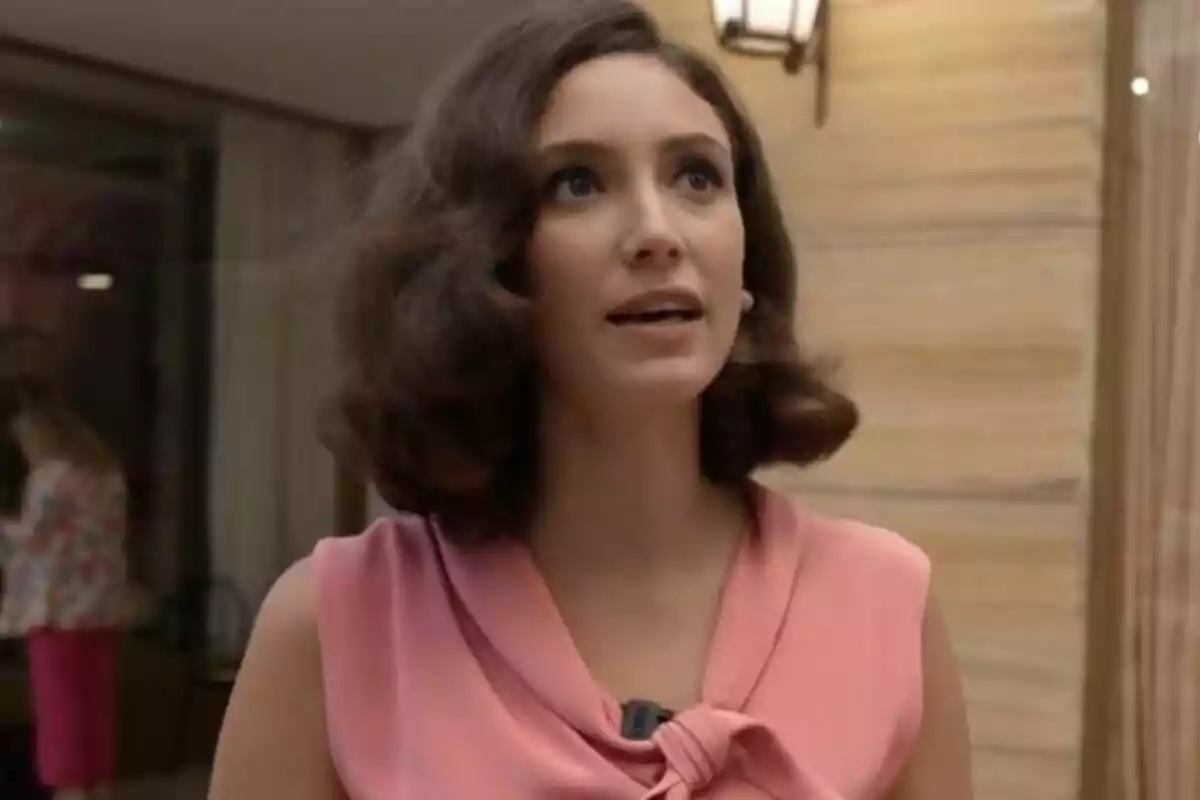 Lucrezia Massari with dark wavy hair, wearing a pink blouse, in an interior setting with wooden walls and a lamp on the wall.