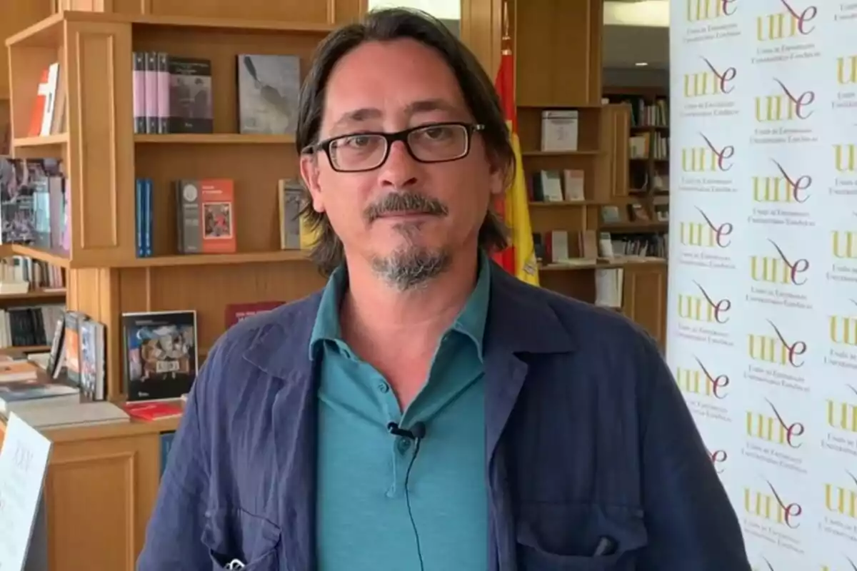 Luis Felipe Torrente amb ulleres i barba està dret en una biblioteca, amb prestatgeries plenes de llibres darrere seu i un cartell de la Unió d'Editorials Universitàries Espanyoles (UNE) a la dreta.