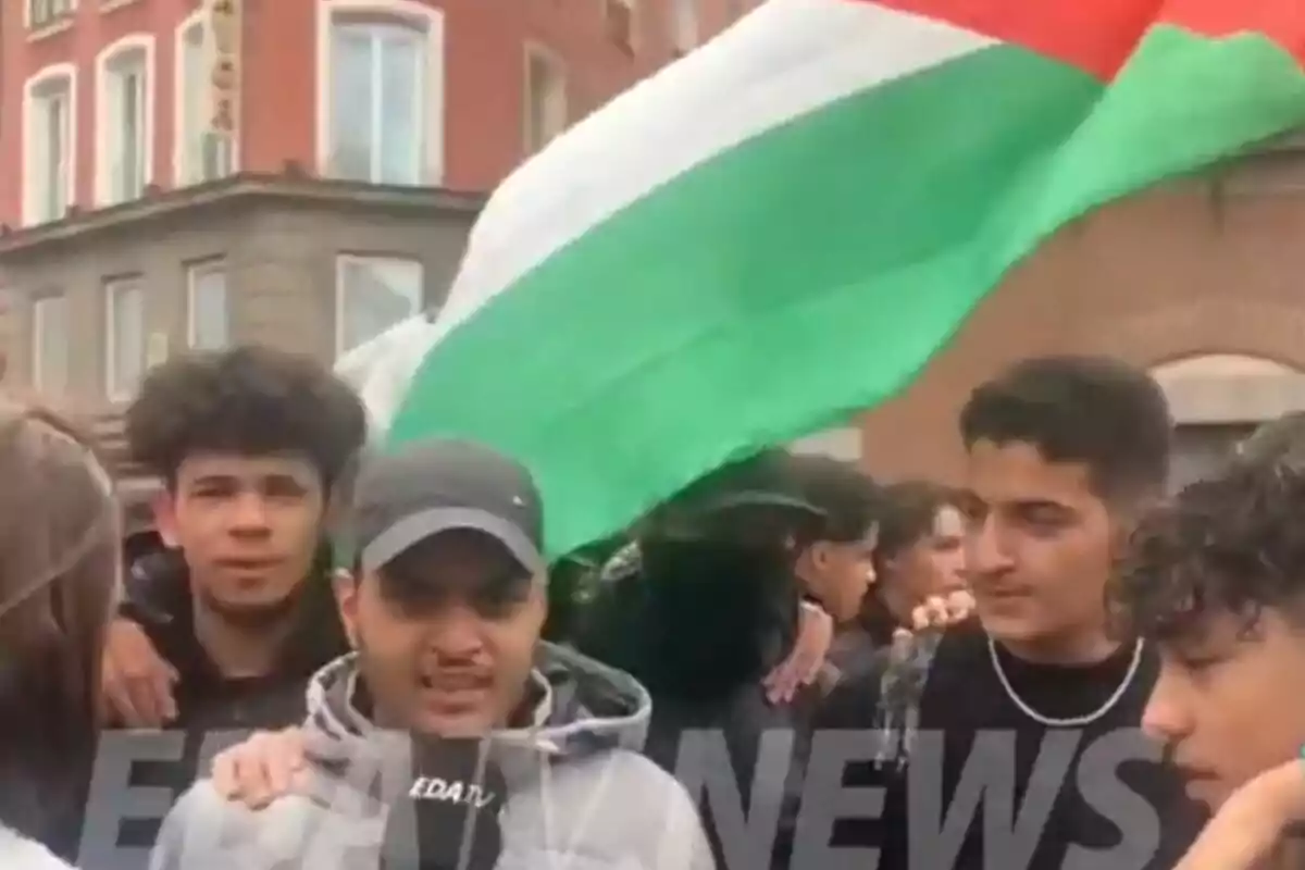 Un grupo de jóvenes se reúne en una calle con una gran bandera en el fondo.