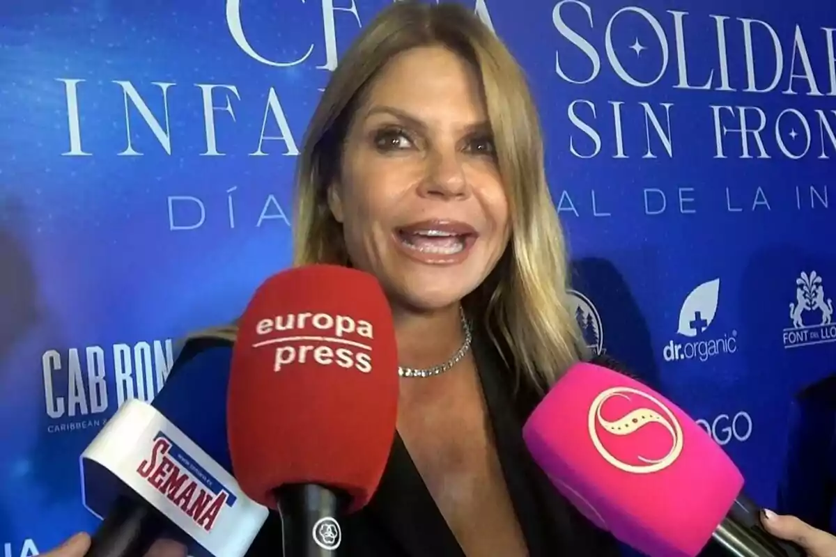 Blonde Makoke smiles as she is interviewed by various media outlets in front of a blue background with logos and text.