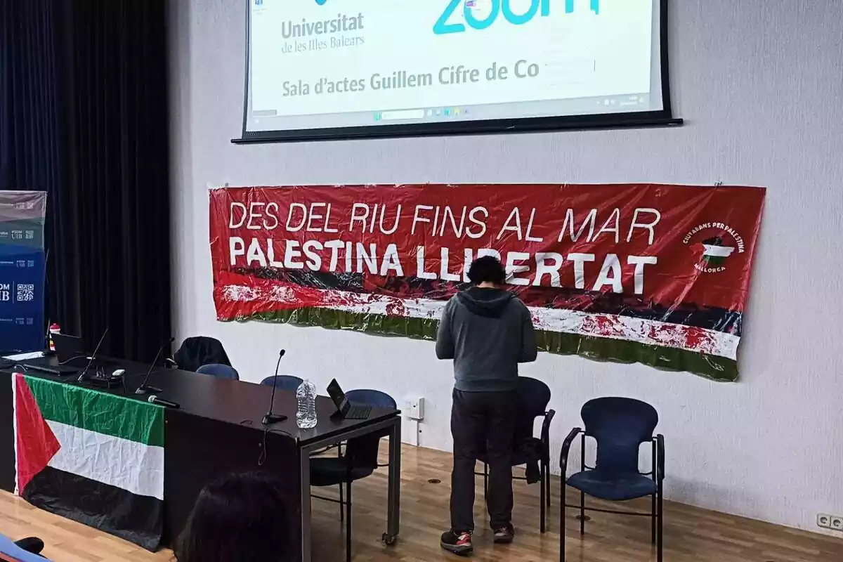 Sala de la Universitat de les Illes Balears amb una bandera palestina a la taula dels conferenciants i de fons un pancarta amb el lema "Palestina llibertat, des del riu fins al mar"