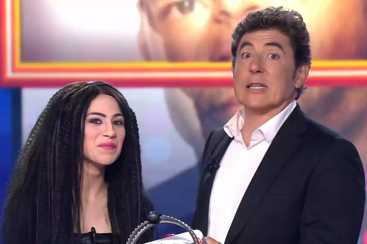 Melani García i Manel Fuentes en un plató de televisió de Tu Cara Me Suena, una dona amb cabell llarg i arrissat i un home amb vestit fosc i camisa blanca, tots dos mirant cap a la càmera.