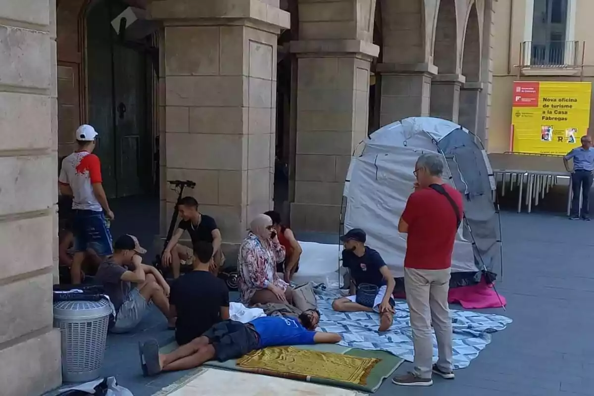 Un grup de persones es troben reunits en una plaça, algunes assegudes i d'altres ajagudes sobre mantes, al costat d'una tenda de campanya i un edifici de pedra amb arcs.