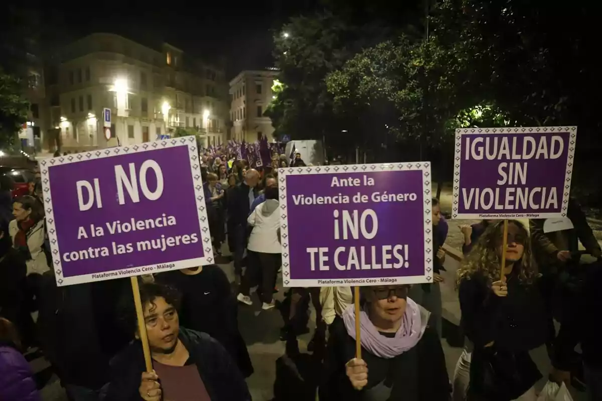 Persones marxant de nit amb pancartes morades que diuen "Digues no a la violència contra les dones", "Davant la violència de gènere no callis" i "Igualtat sense violència".