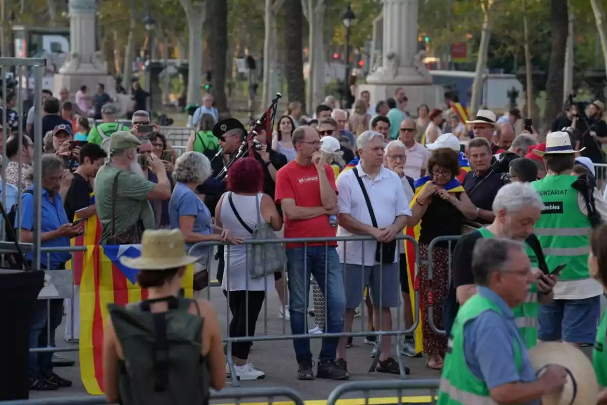 Un grup de persones es troben reunits en un espai públic, algunes portant banderes i altres fent fotos, mentre un home toca la gaita i diversos voluntaris amb armilles verdes són presents.