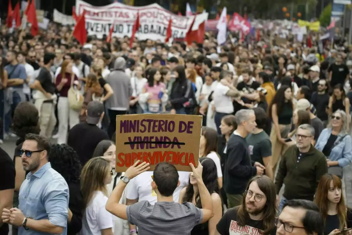 Una multitud de persones participa en una manifestació, algunes porten pancartes i banderes, mentre un individu sosté un cartell que diu “Ministeri de Supervivència”.