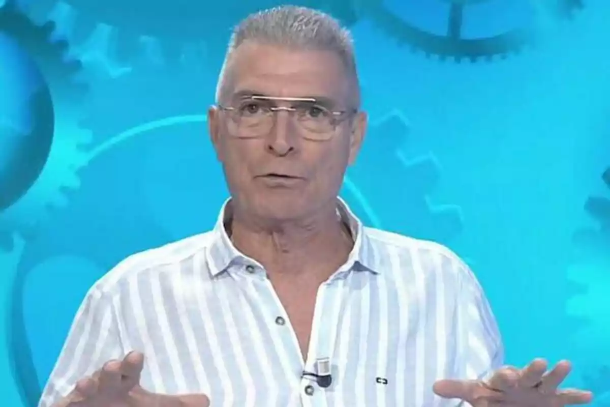 Manolo Sarria amb camisa de ratlles i ulleres parla davant d'un fons blau amb engranatges a Atrápame si pots.