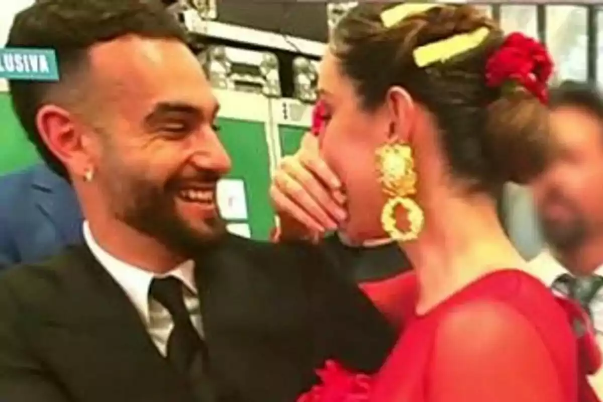 Manuel Cortés i Adara Molinero somrient comparteixen un moment d'alegria, amb la dona vestint un vestit vermell i arracades daurades, mentre l'home porta un vestit fosc.