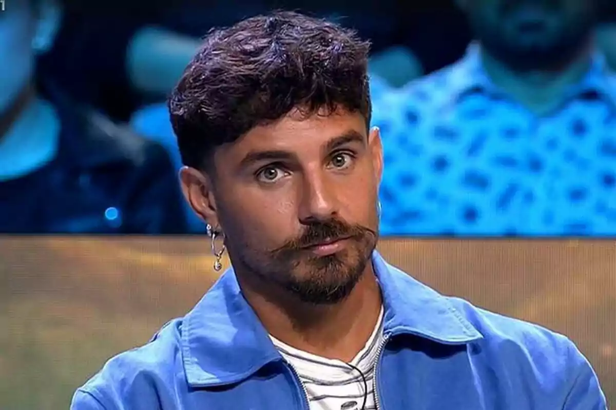 Manuel González amb barba i bigoti porta una jaqueta blava i una arracada a l'orella mentre està assegut en un entorn d'estudi al plató de Supervivientes amb llums blaves de fons.
