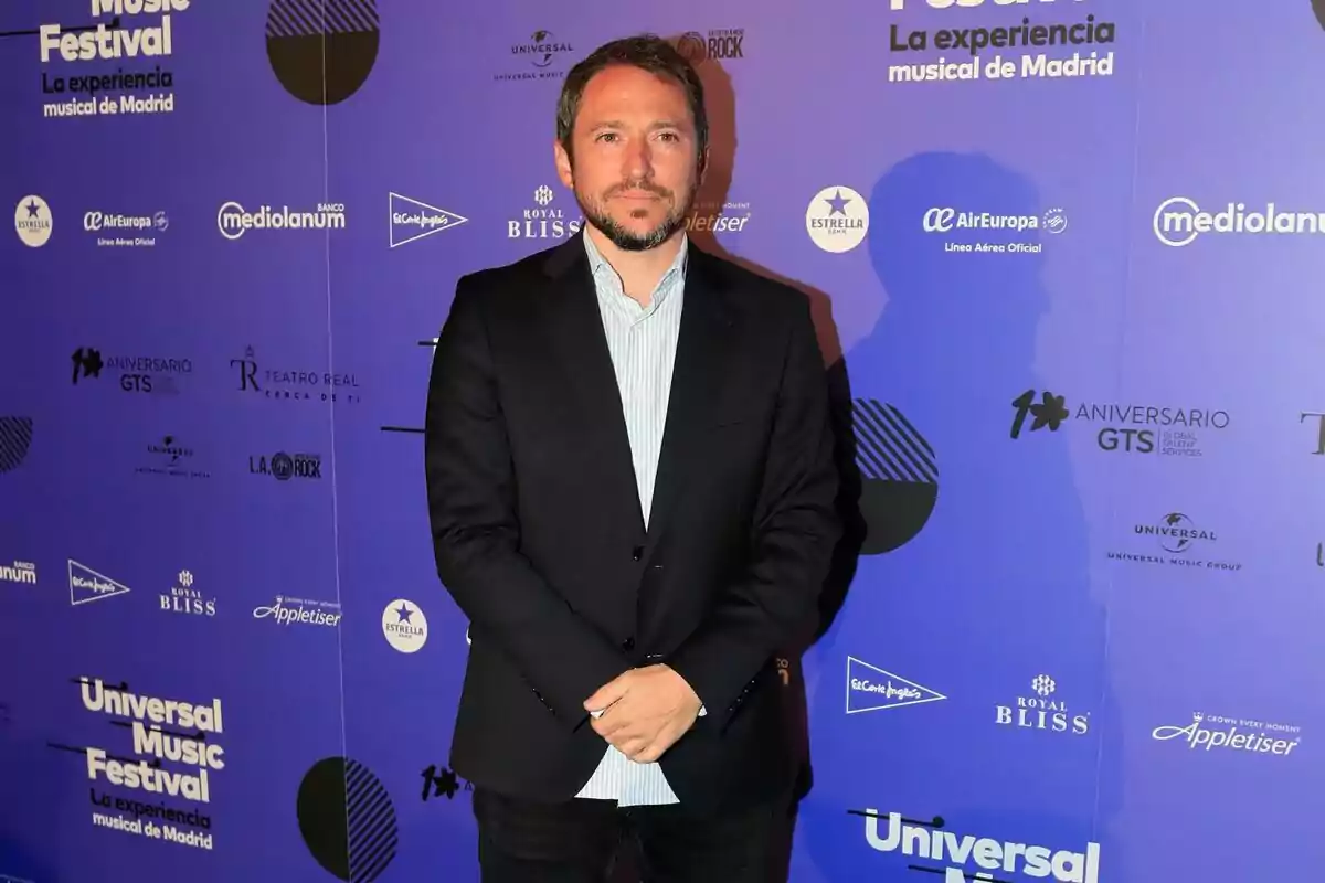 Manuel Martos con traje oscuro y camisa clara posa frente a un fondo azul con logotipos de patrocinadores en un evento musical en Madrid.