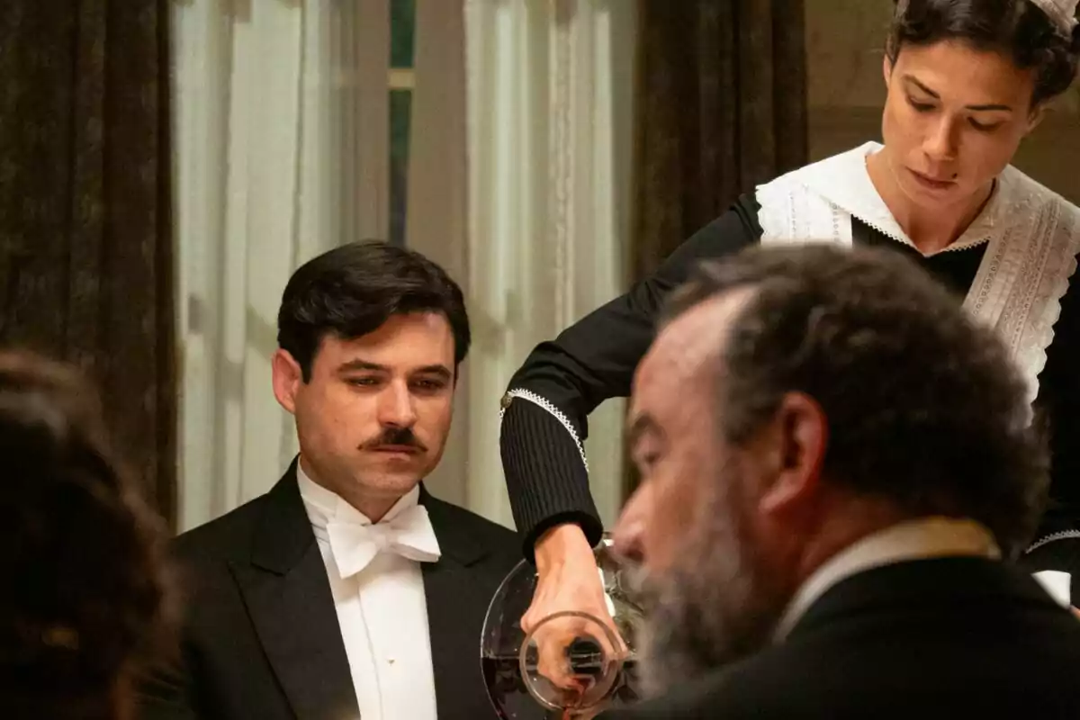 Una cambrera serveix vi a Manuel amb bigoti que està assegut en una taula elegantment vestida, mentre que un altre home de barba està en primer pla a La Promesa.