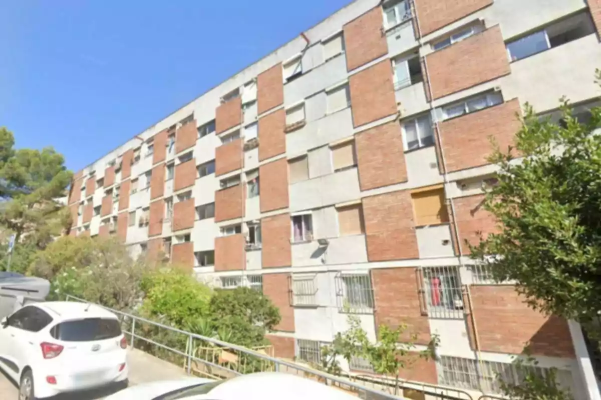 Edifici residencial de diversos pisos amb façana de maó i ciment, envoltat d’arbres i cotxes aparcats al carrer. Edifici residencial de diversos pisos amb façana de maó i ciment, envoltat d’arbres i cotxes aparcats al carrer.