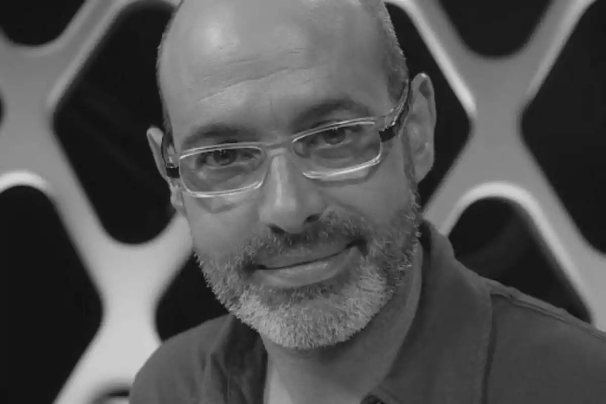 Fotografía en blanco y negro de Marc Boada con gafas y barba canosa sonriendo frente a un fondo con patrón geométrico.