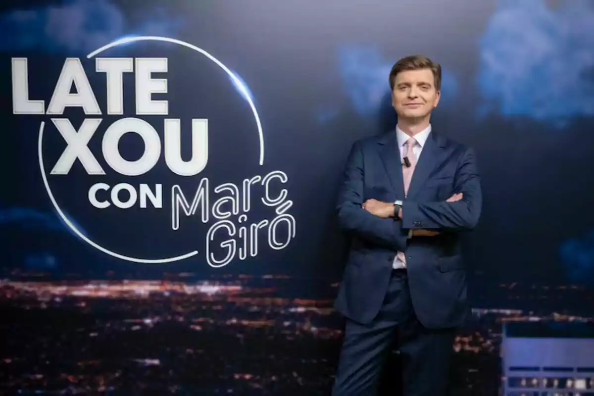 Marc Giró con un traje posa con los brazos cruzados frente a un fondo que publicita su programa, 