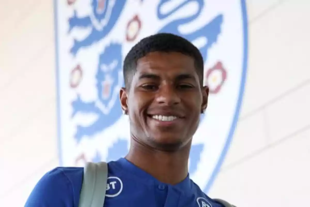 A smiling man with a blue t-jersey and a backpack, in front of a shield with blue lions. A smiling man with a blue t-jersey and a backpack, in front of a shield with blue lions.