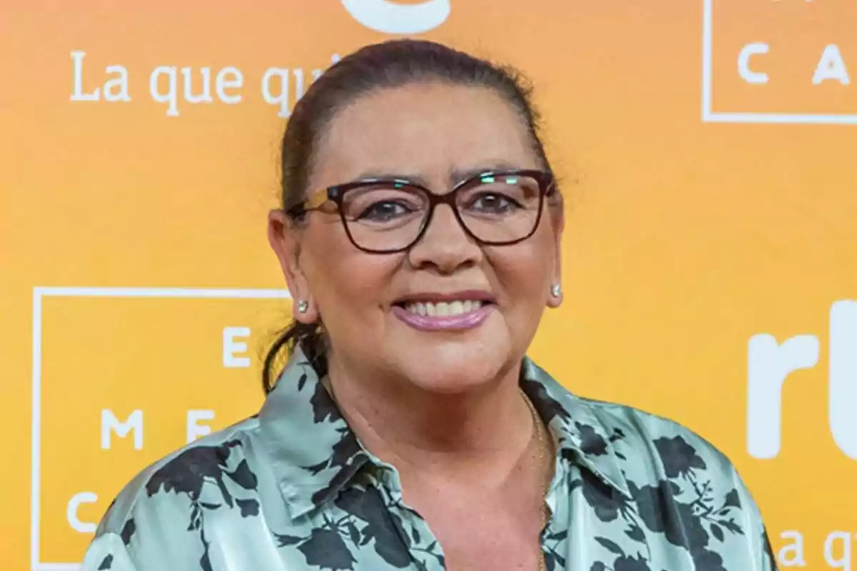 María del Monte amb ulleres i camisa estampada somriu davant d'un fons taronja amb text en un photocall de RTVE.