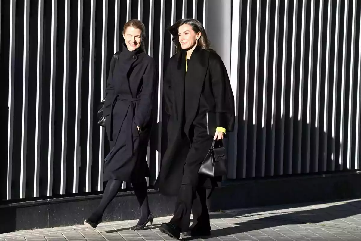 María Dolores Ocaña i la reina Letícia caminant juntes, vestides amb abrics foscos, davant d'una paret amb barres verticals. María Dolores Ocaña i la reina Letícia caminant juntes, vestides amb abrics foscos, davant d'una paret amb barres verticals.