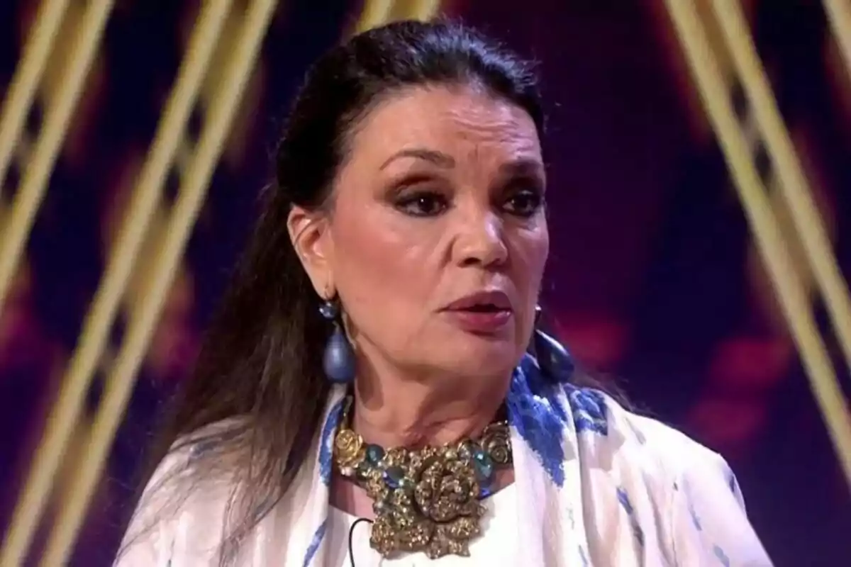 María José Cantudo amb cabell fosc i solt, portant un collaret gran i arracades, està en un escenari amb llums de fons a De Viernes. María José Cantudo amb cabell fosc i solt, portant un collaret gran i arracades, està en un escenari amb llums de fons a De Viernes.