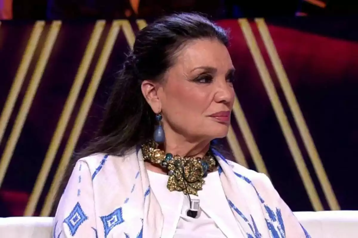María José Cantudo amb cabell fosc i recollit porta un collaret daurat i blau i una arracada blava, vestida amb una peça blanca amb detalls blaus, asseguda en un entorn il·luminat a De Viernes. María José Cantudo amb cabell fosc i recollit porta un collaret daurat i blau i una arracada blava, vestida amb una peça blanca amb detalls blaus, asseguda en un entorn il·luminat a De Viernes.
