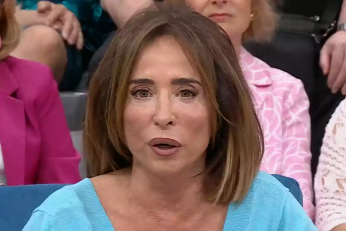 María Patiño amb cabell castany i expressió seriosa està asseguda en un entorn d'estudi de televisió a La Família de la Tele, envoltada d'altres persones.
