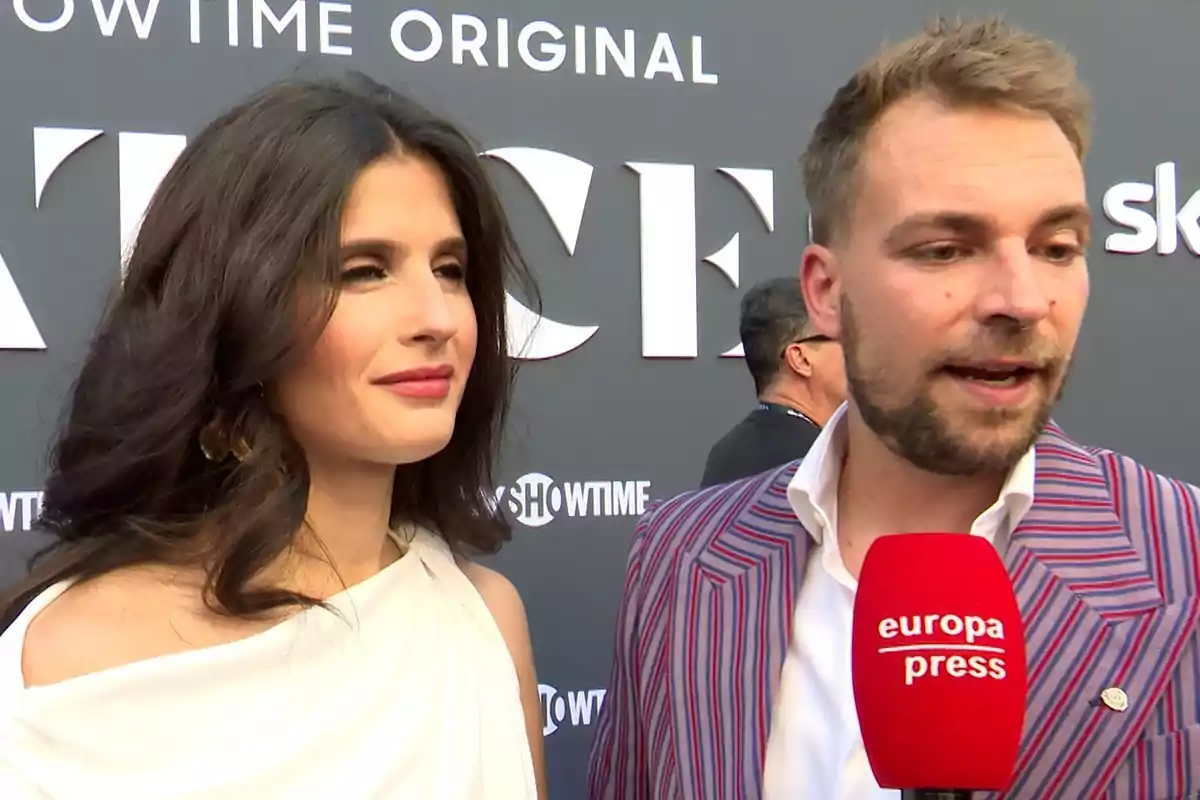 María Sánchez in a white dress and José María Almoguera in a striped jacket are speaking in front of a red Europa Press microphone at an event with a background of large letters. María Sánchez in a white dress and José María Almoguera in a striped jacket are speaking in front of a red Europa Press microphone at an event with a background of large letters.
