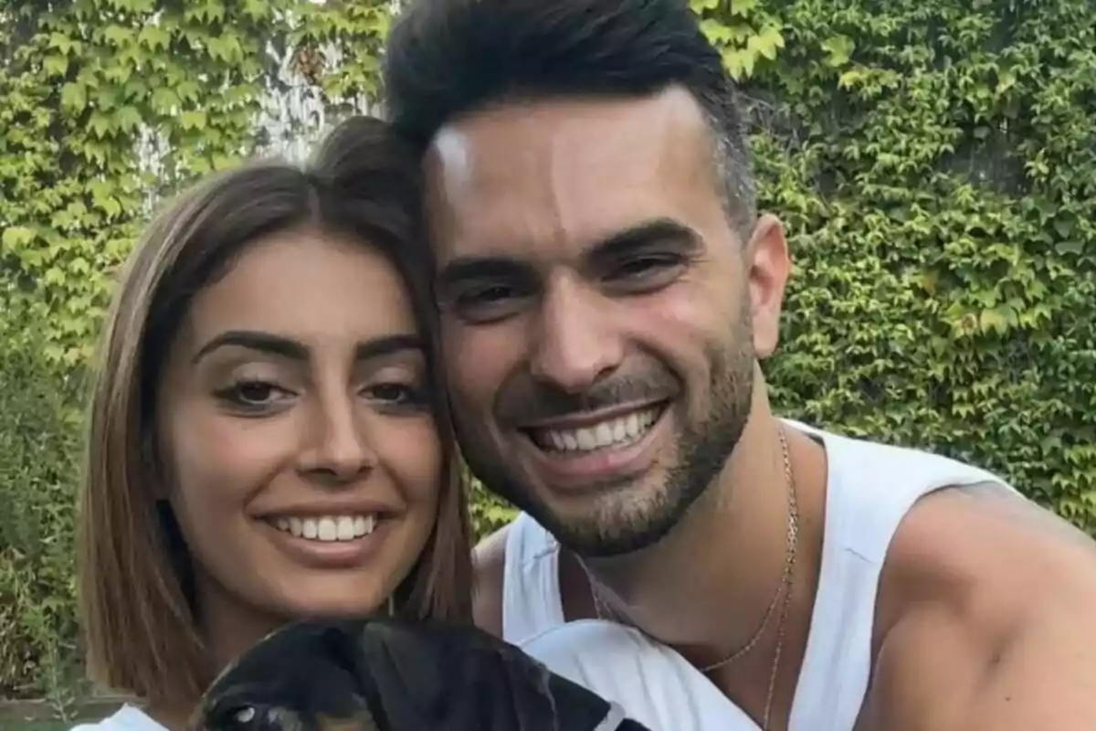 Capture of Marieta Díaz and Suso Álvarez smiling posing with a dog in front of a background of vegetation.