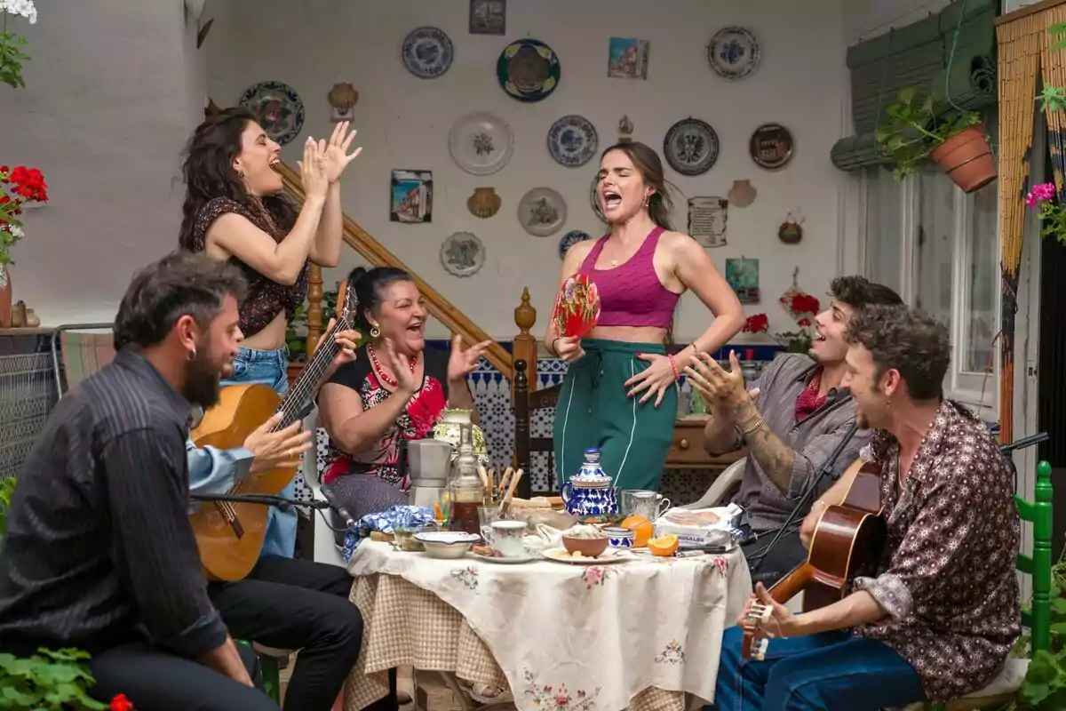 Un grup de persones amb la Melody com a protagonista gaudeix d’una trobada animada en un pati decorat amb plats a la paret, mentre toquen la guitarra, canten i aplaudeixen al voltant d’una taula amb menjar i beguda a Mariliendre. Un grup de persones amb la Melody com a protagonista gaudeix d’una trobada animada en un pati decorat amb plats a la paret, mentre toquen la guitarra, canten i aplaudeixen al voltant d’una taula amb menjar i beguda a Mariliendre.