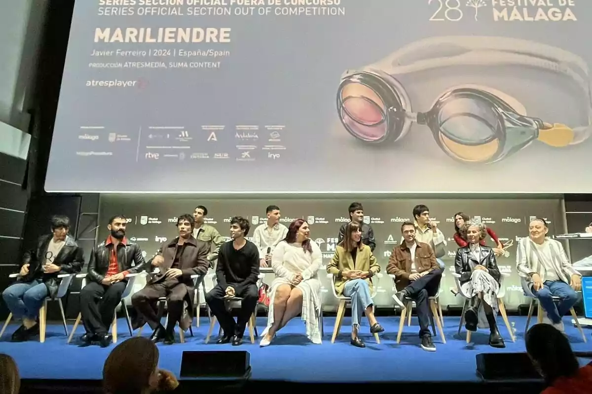 Un grupo de personas está sentado en un escenario durante un evento del Festival de Málaga, con un cartel de fondo que anuncia una serie llamada "Mariliendre".