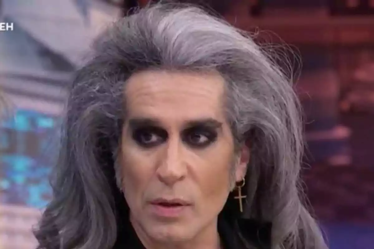 Mario Vaquerizo con cabello largo y canoso, maquillaje oscuro alrededor de los ojos y un pendiente en forma de cruz en 'El Hormiguero'.