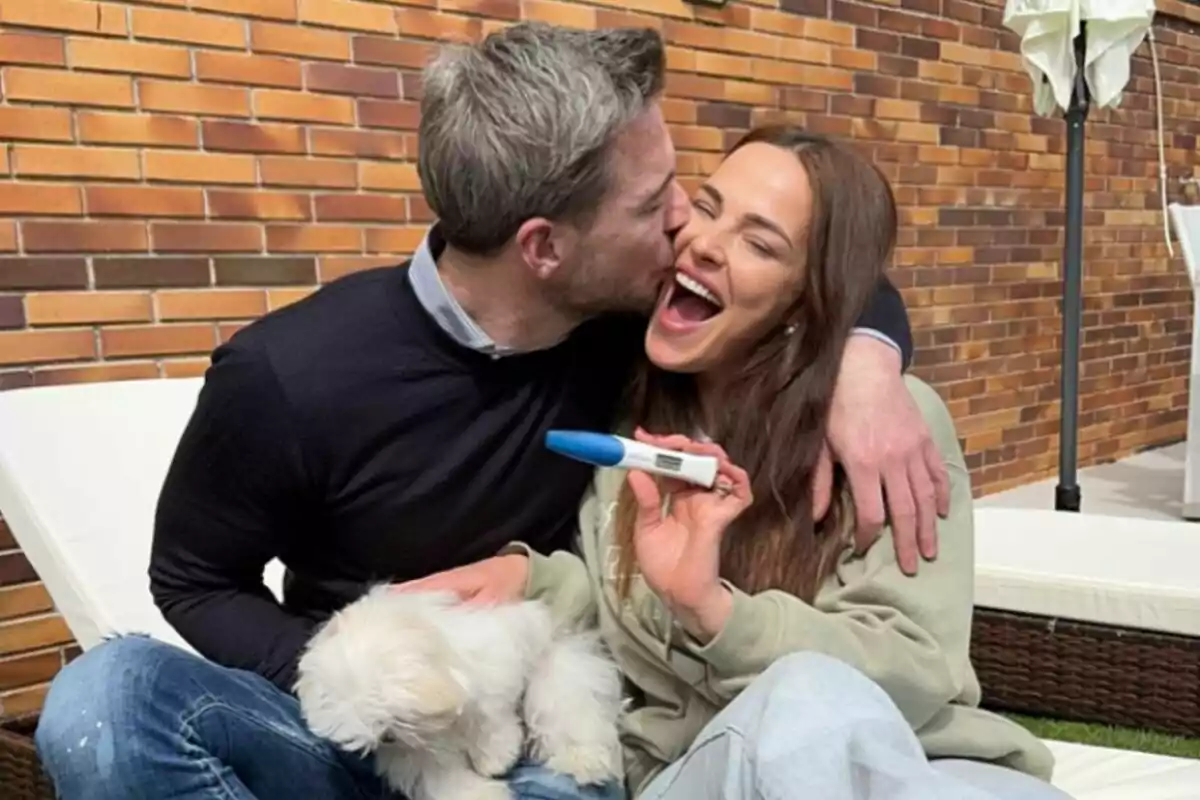 Marta Peñate i Tony Spina somrients s'abracen mentre la dona sosté una prova d'embaràs positiva i un petit gos blanc està a la seva falda. Marta Peñate i Tony Spina somrients s'abracen mentre la dona sosté una prova d'embaràs positiva i un petit gos blanc està a la seva falda.