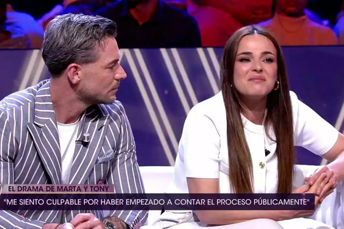 Tony Spina amb jaqueta de ratlles i Marta Peñate de blanc estan asseguts en un plató de televisió de De Divendres amb públic al fons i un rètol a la pantalla que esmenta el drama de la Marta i en Tony. Tony Spina amb jaqueta de ratlles i Marta Peñate de blanc estan asseguts en un plató de televisió de De Divendres amb públic al fons i un rètol a la pantalla que esmenta el drama de la Marta i en Tony.
