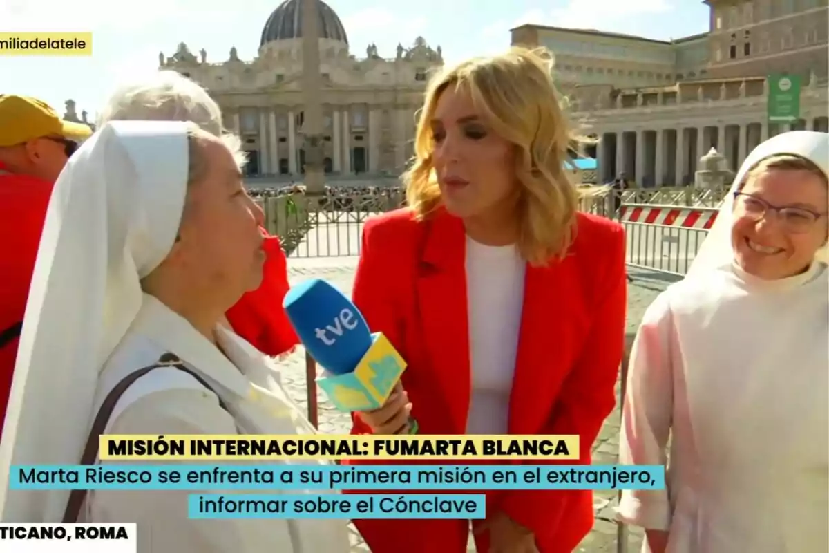 Marta Riesco amb un micròfon de TVE a La Família de la Tele entrevista una monja a la Plaça de Sant Pere al Vaticà mentre una altra monja somriu al costat. Marta Riesco amb un micròfon de TVE a La Família de la Tele entrevista una monja a la Plaça de Sant Pere al Vaticà mentre una altra monja somriu al costat.