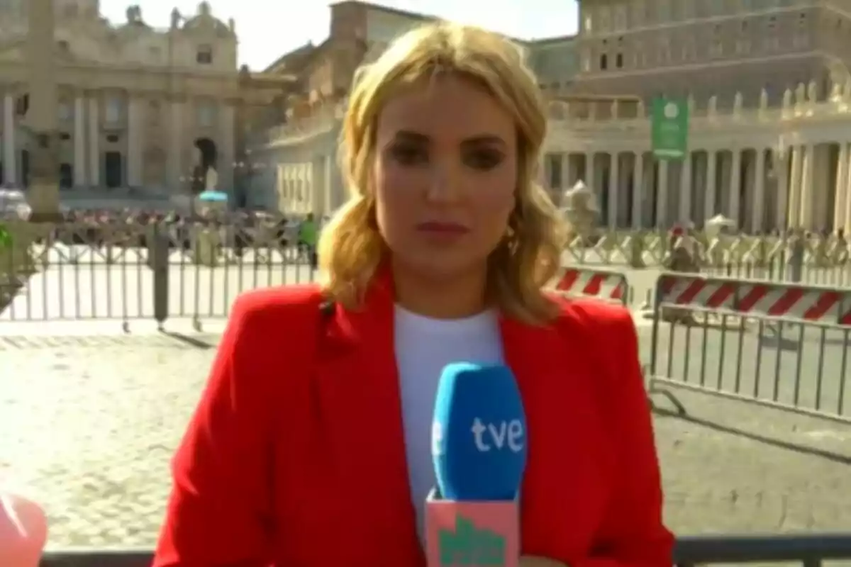 Marta Riesco amb jaqueta vermella sosté un micròfon de TVE a La Família de la Tele davant d'un edifici històric amb columnes i una plaça concorreguda.
