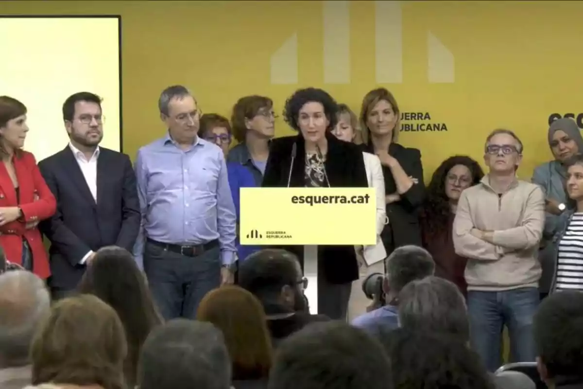 Un grup de persones en un esdeveniment d?Esquerra Republicana amb una dona parlant en un podi groc davant d?una audiència.