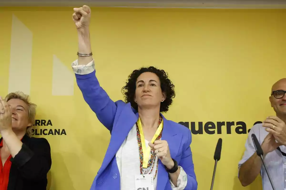 Marta Rovira d?ERC aixeca el puny en un esdeveniment amb un fons groc que té text parcialment visible.