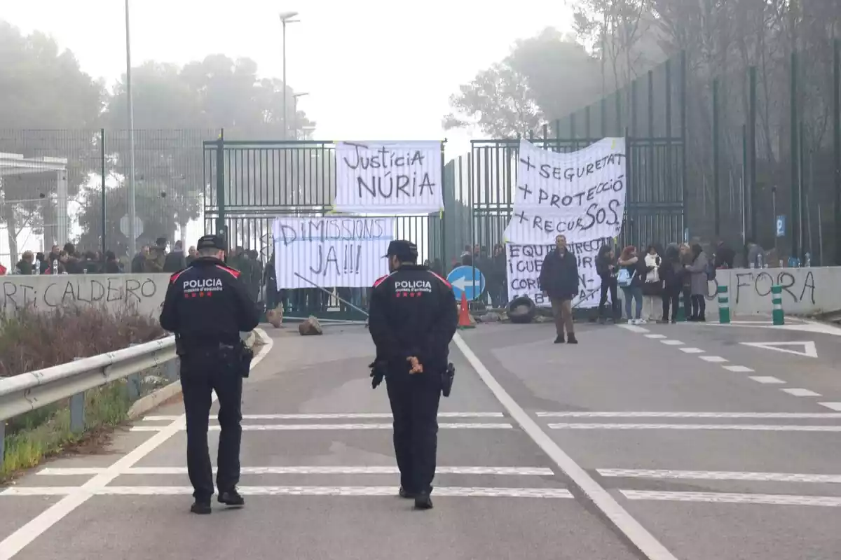 Dos Mossos d'Esquadra d'esquena davant de desenes de treballadors a les portes de la presó de Mas d'Enric amb pancartes demanant justícia per a la Núria, dimissions i més mesures de seguretat