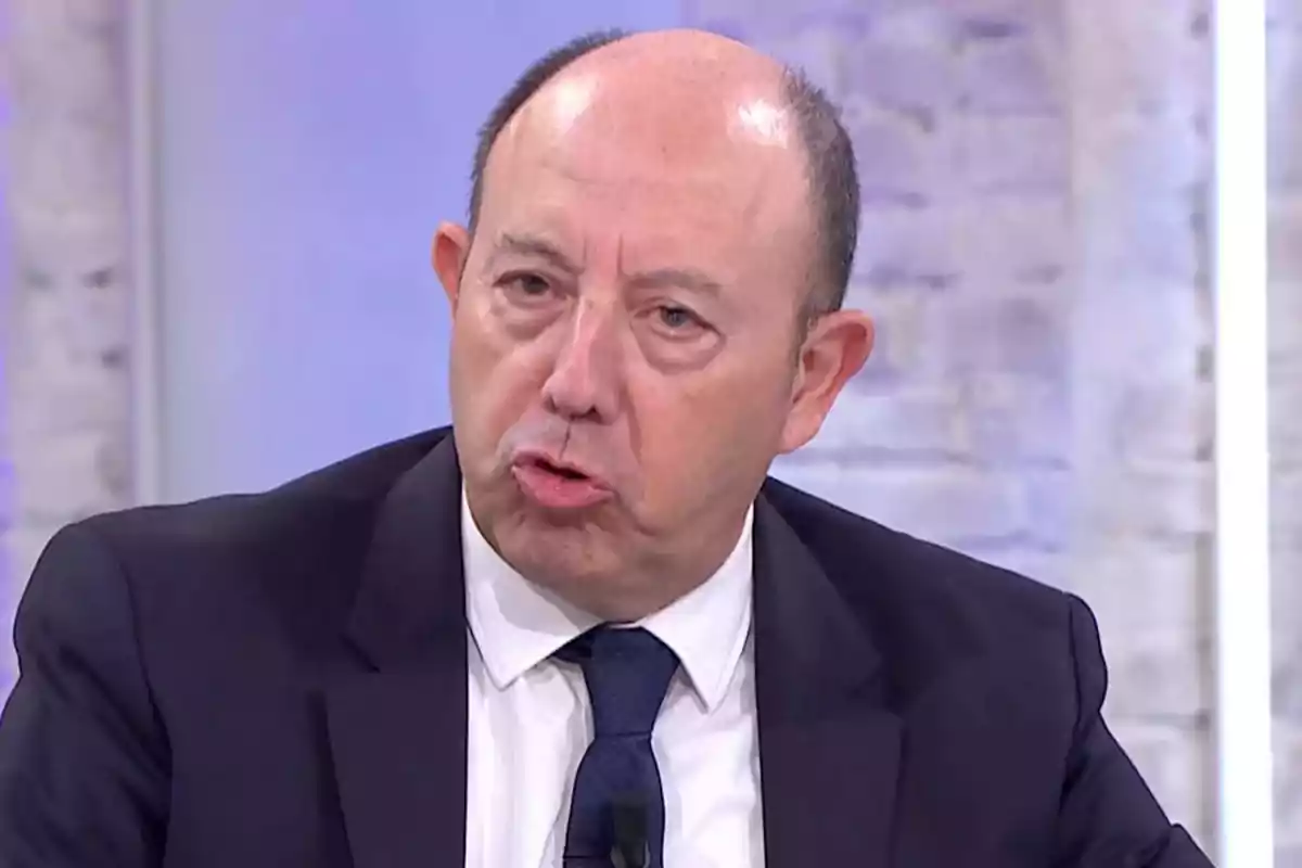 Home calb de mitjana edat amb vestit fosc i corbata blava parlant en un estudi de televisió amb fons clar Home calb de mitjana edat amb vestit fosc i corbata blava parlant en un estudi de televisió amb fons clar