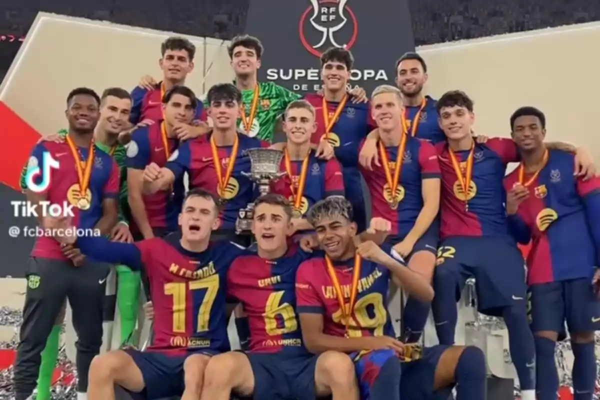 Jugadors del Barcelona celebren amb medalles i un trofeu després de guanyar la Supercopa d’Espanya posant somrients davant d’un cartell del torneig Jugadors del Barcelona celebren amb medalles i un trofeu després de guanyar la Supercopa d’Espanya posant somrients davant d’un cartell del torneig