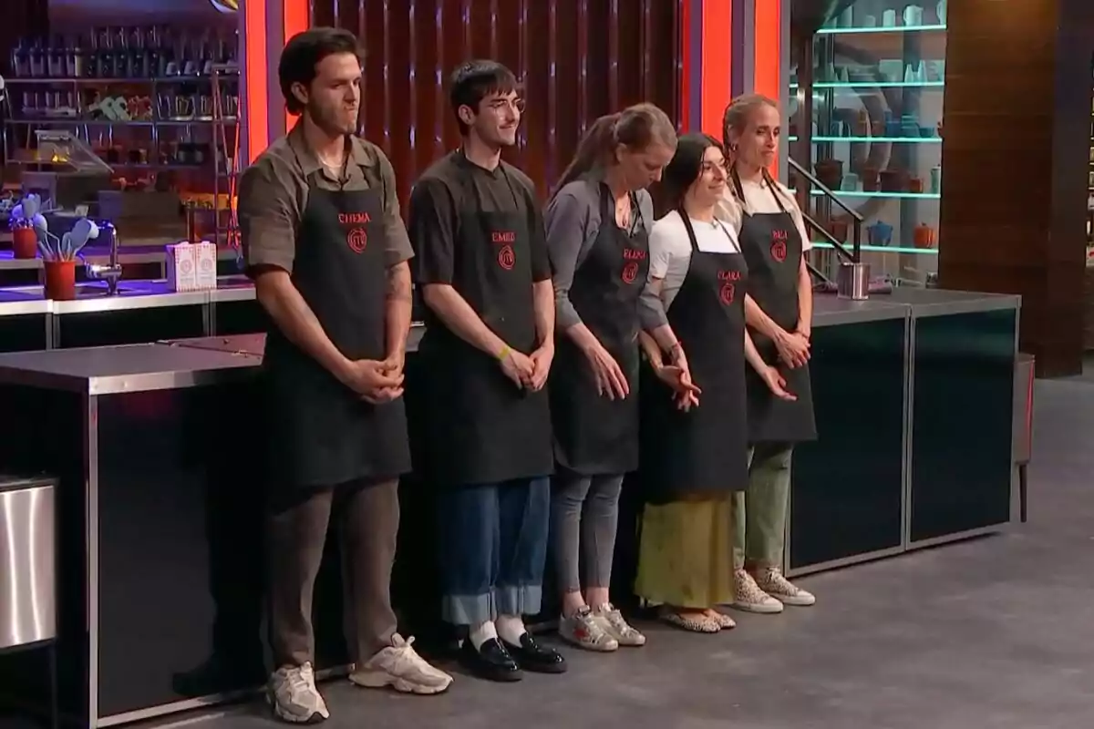 Cinc concursants de 'Masterchef' estan drets en fila en una cuina professional, tots porten davantals negres amb els seus noms i el logotip del programa.