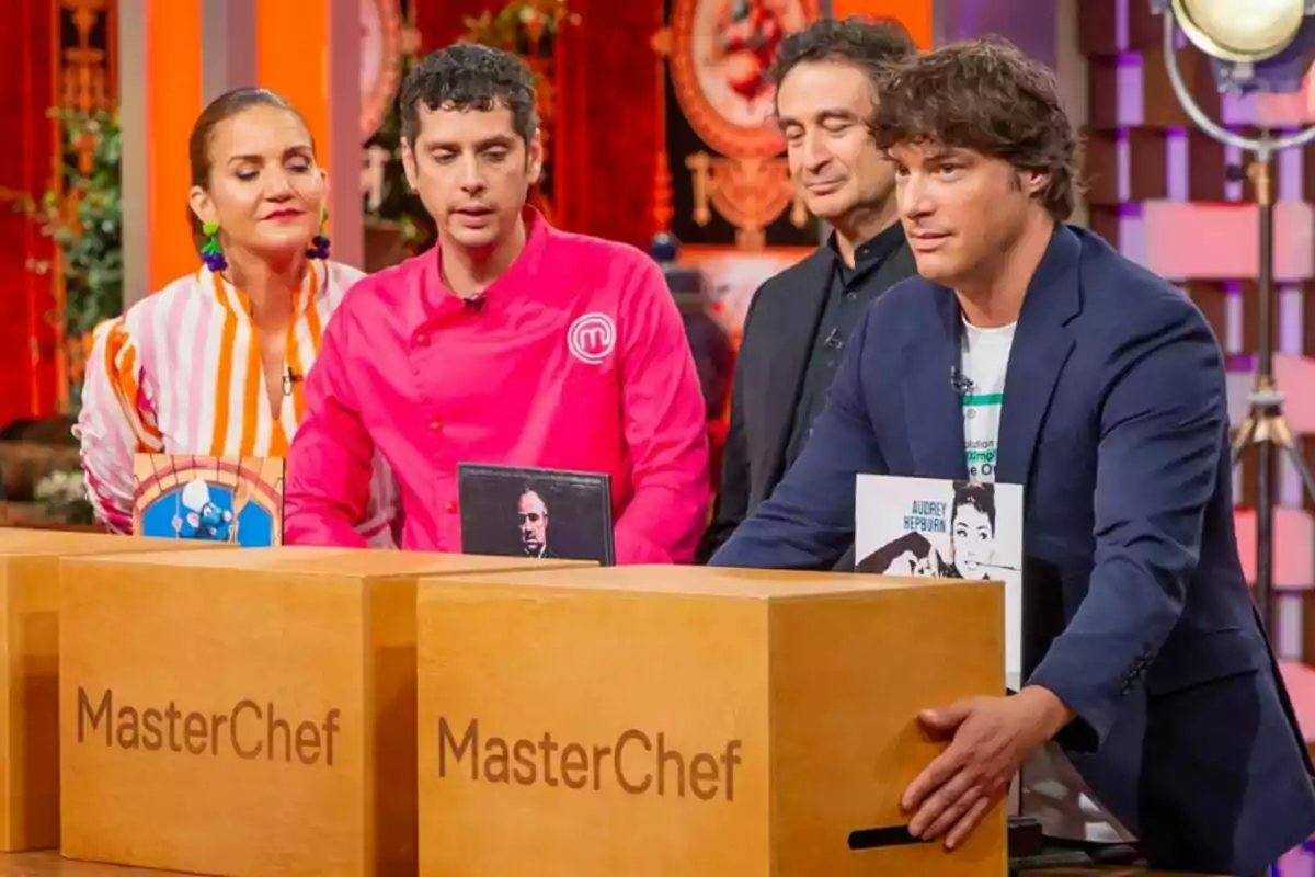 Jordi Cruz, Pepe Rodríguez i Samantha Vallejo Nágera amb Eduardo Casanova estan drets darrere de caixes de fusta amb el logotip de MasterChef en un set de televisió colorit.