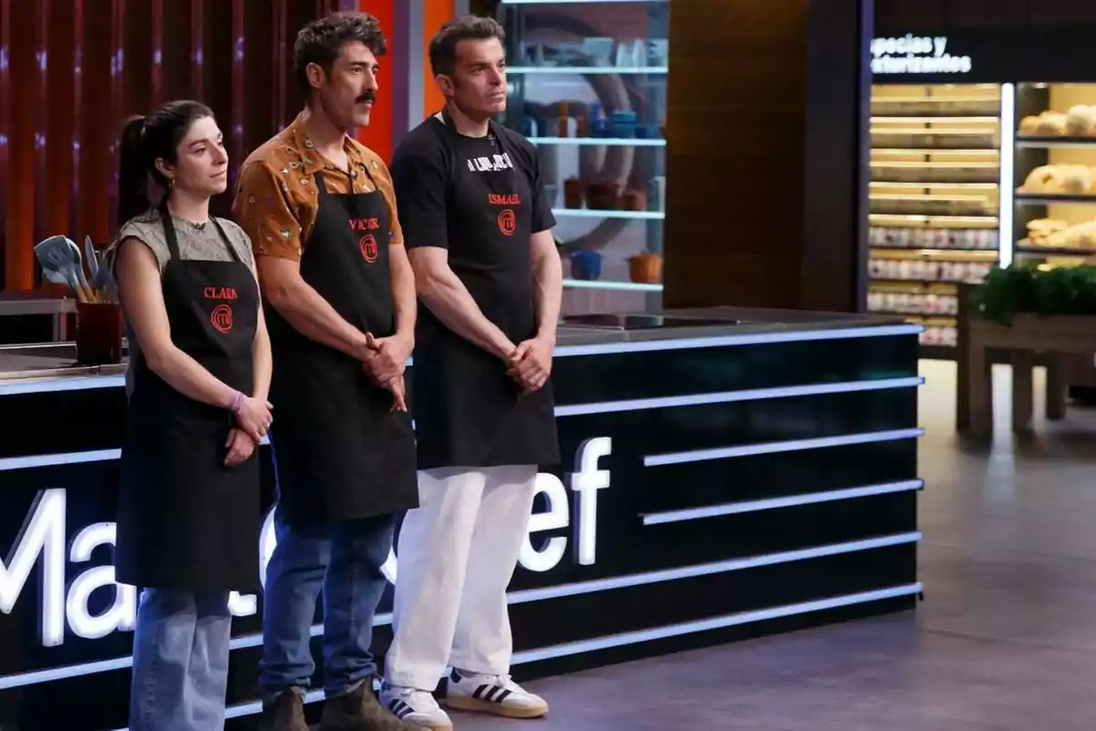 Tres persones amb davantals negres estan dretes en un plató de cuina, participant a 'Masterchef'.