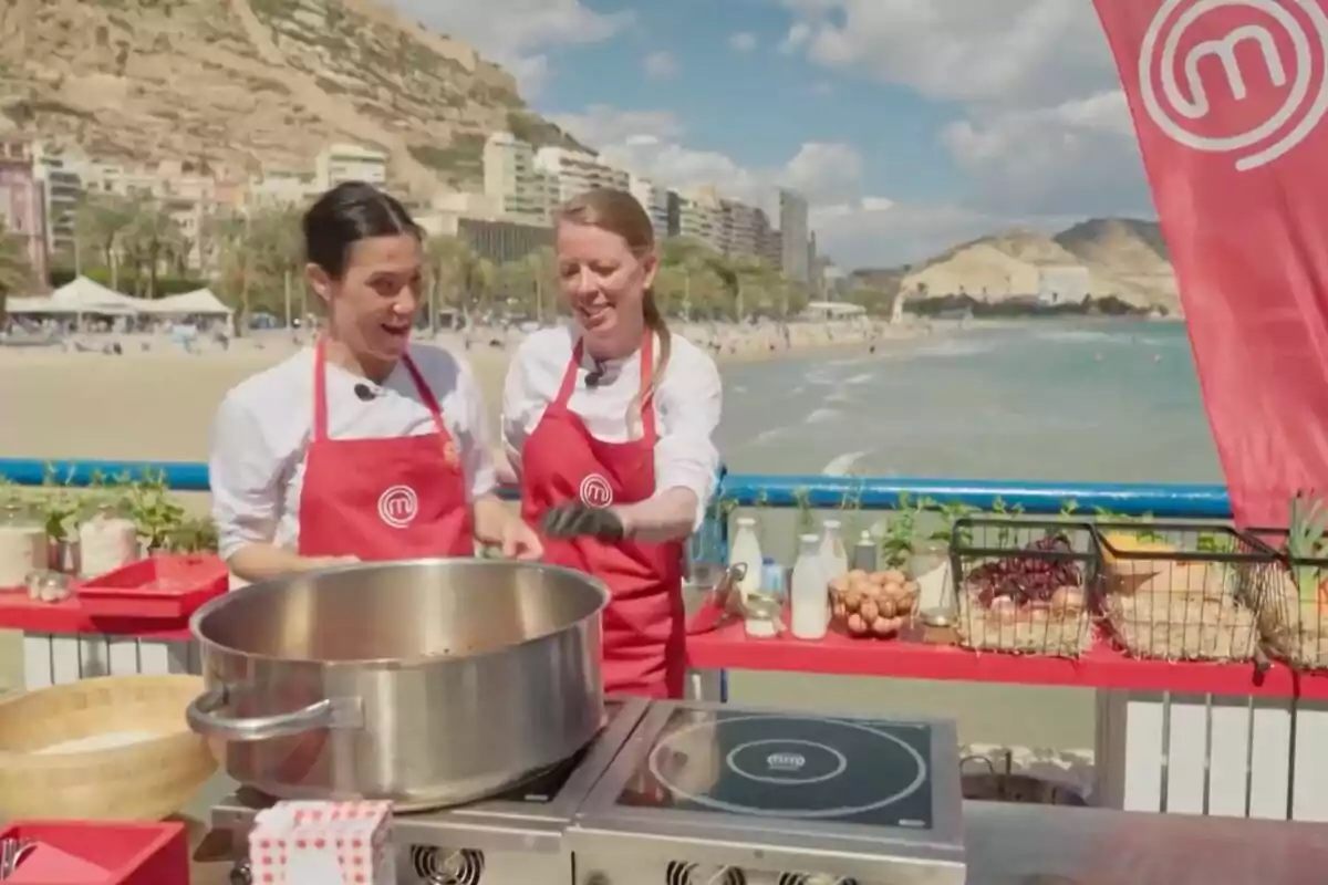 Dues persones amb davantals vermells cuinen a l’aire lliure al costat de la platja, amb ingredients i utensilis sobre una taula i edificis i muntanyes al fons a ‘MasterChef’. Dues persones amb davantals vermells cuinen a l’aire lliure al costat de la platja, amb ingredients i utensilis sobre una taula i edificis i muntanyes al fons a ‘MasterChef’.