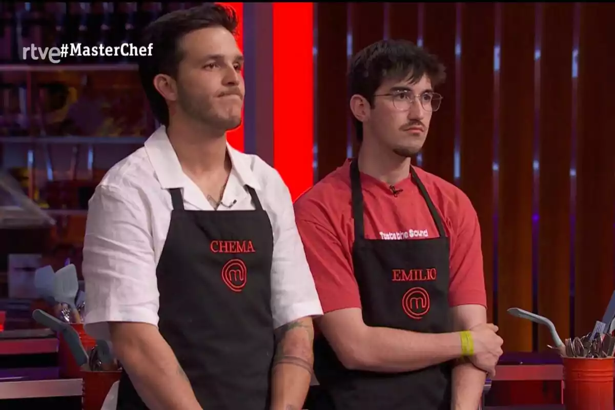 Dos concursants de MasterChef amb davantals negres i noms brodats en vermell estan drets a la cuina del programa amb una expressió seriosa.