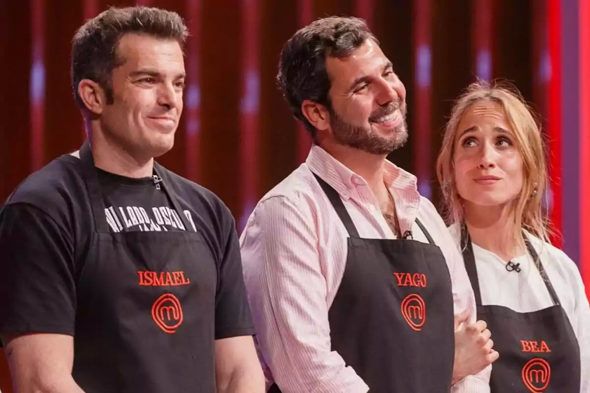 Tres concursants de MasterChef amb davantals negres i noms brodats posen junts al plató del programa amb expressions d’expectativa i emoció.