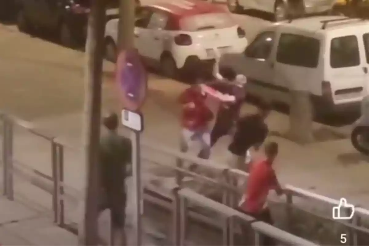 Diverses persones corren i semblen estar en una confrontació en un carrer amb cotxes estacionats.