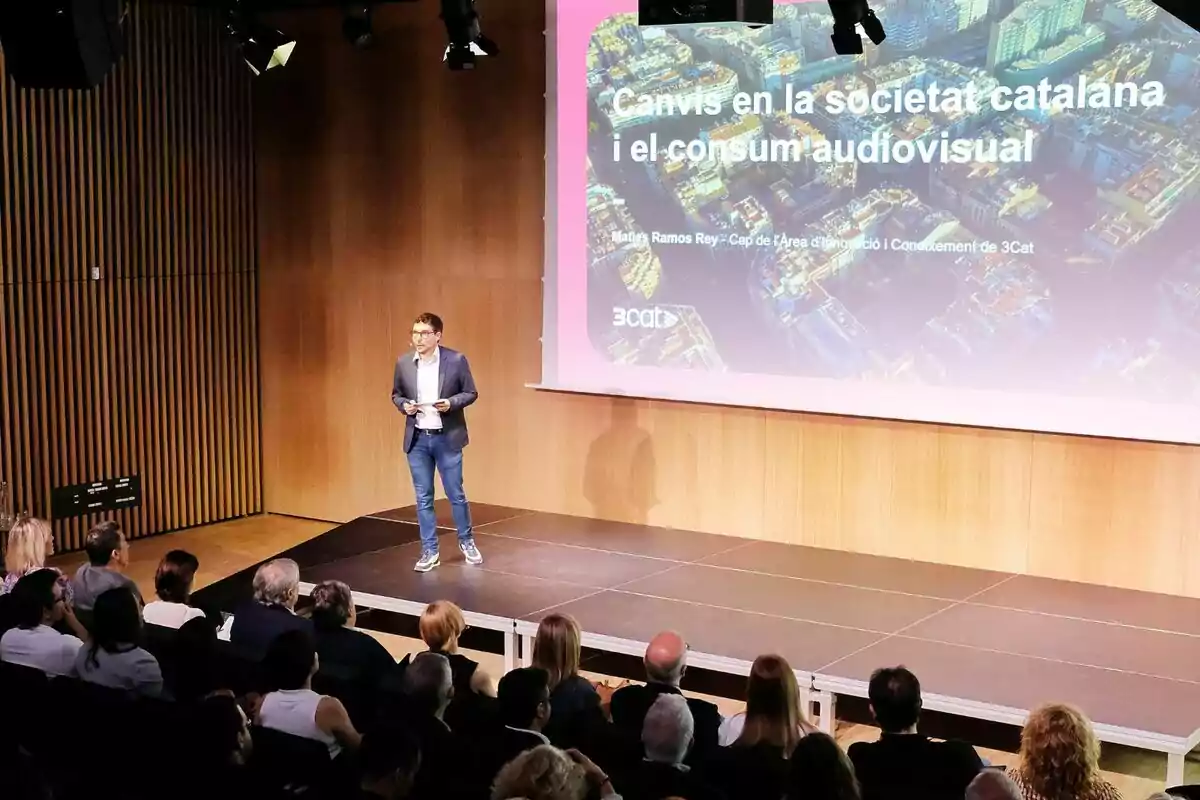 Maties Ramos dret damunt d’un escenari fa una presentació davant d’una audiència, amb una pantalla gran al fons que mostra un títol sobre els canvis en la societat catalana i el consum audiovisual de 3Cat. Maties Ramos dret damunt d’un escenari fa una presentació davant d’una audiència, amb una pantalla gran al fons que mostra un títol sobre els canvis en la societat catalana i el consum audiovisual de 3Cat.