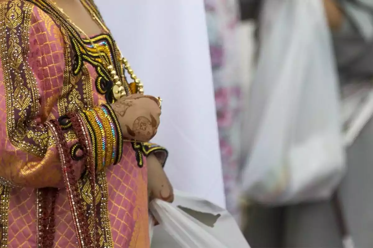 Person in colorful traditional attire with golden details, holding a plastic bag, with henna designs on their hand.