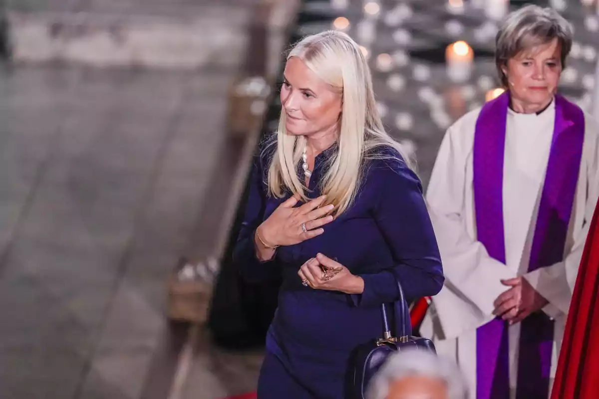 Mette-Marit in a dark blue dress stands in a religious setting, with one hand on her chest and a solemn expression, while another person in clerical attire is behind her. Mette-Marit in a dark blue dress stands in a religious setting, with one hand on her chest and a solemn expression, while another person in clerical attire is behind her.
